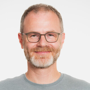 Mann mit Brille, Bart und Lächeln vor hellem Hintergrund.